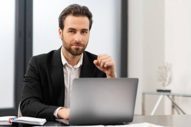 Erkek girişimcinin kameraya bakmasına sevindim. Resmi kıyafetler giyen neşeli beyaz adam masada oturuyor ve ofiste dizüstü bilgisayar kullanıyor.