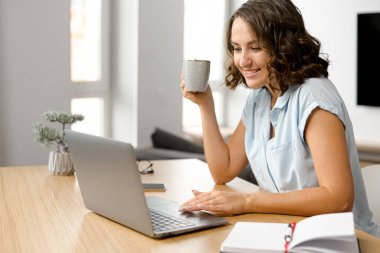 Serbest çalışan bir kadın bir fincan sabah kahvesiyle hayal kuruyor, gülümseyen bir kadın elinde dizüstü bilgisayarla masaya oturuyor ve ekrana bakıyor.