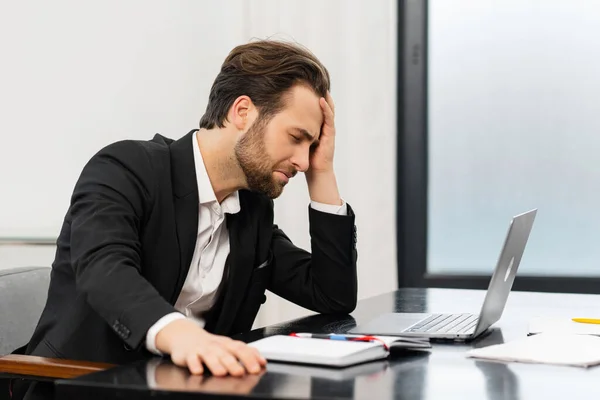 Homem de negócios cansado franzir a testa enquanto verifica documentos financeiros, tocando na testa com o braço, sentado no escritório da moda no laptop — Fotografia de Stock