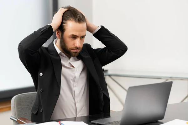 Frustrated caucasian unshaved entrepreneur with sad grimace in front of laptop in office, noticed a big mistake he did, touches his head with his hand — Stock Photo, Image