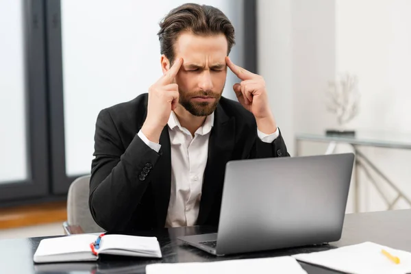 Tired businessman frown while checking financial documents, touching forehead with the arm, sitting in the trendy office at the laptop, consulting client online, analyze statistic — Stock Photo, Image