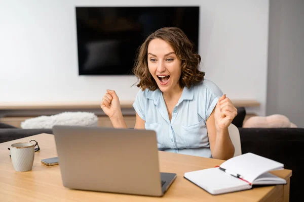 Dosažení cíle. Veselá rozradostněná mladá žena křičí ano a zvedá ruce v gestu vítězství, sedí před laptopem v domácí kanceláři — Stock fotografie