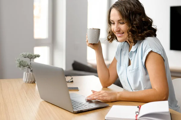 Mujer freelancer soñando despierto con una taza de café de la mañana, mujer sonriente se sienta en la mesa con un ordenador portátil, mira a la pantalla —  Fotos de Stock