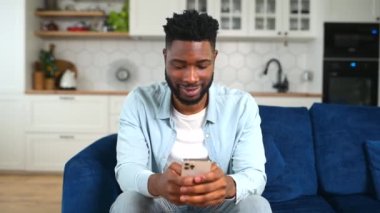 Gülümseyen Afro-Amerikan adam kanepede oturuyor, cep telefonunu sosyal ağları kontrol etmek için kullanıyor.
