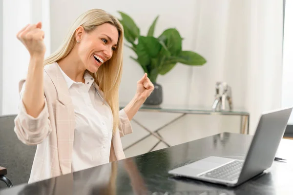 Mladá žena v neformální košili se raduje z úspěchu práce, úspěšné dohody a růstu zisku. Dívá se na monitor notebooku — Stock fotografie