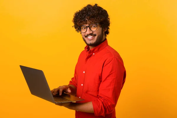Portrét mladého multiraciálního chlapa stojícího izolovaně na oranžové stěně a držícího notebook, píšícího vzkazy, chatujícího s přítelem na sociálních sítích online, sledujícího video — Stock fotografie