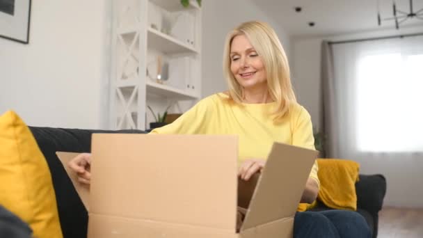 Elegante mujer rubia de mediana edad sentada en el sofá con caja de cartón en las vueltas — Vídeos de Stock