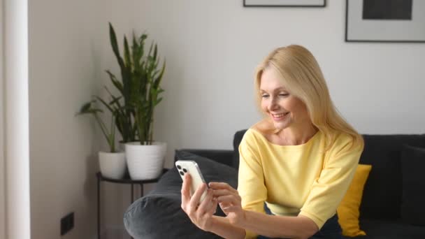 Leende medelålders kvinna med en smartphone sittandes i soffan hemma — Stockvideo