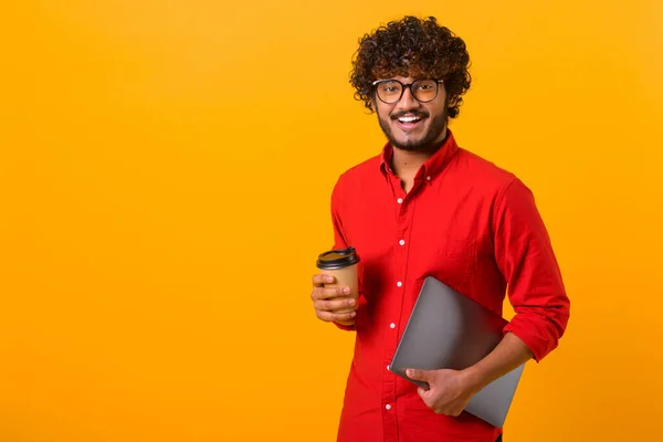 Waist up photo of handsome cheerful smiling Indian man extends hand, holds takeaway coffee cup, drinks coffee or tea, smiling to the camera