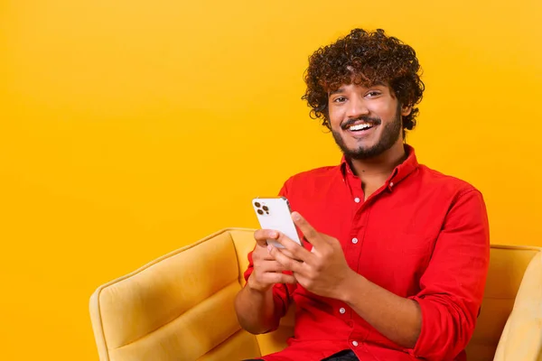 Счастливый индиец смотрит в камеру, прокручивает и печатает сообщение или информационный контент, используя смартфон для чата, сидя в кресле изолированы на оранжевом фоне — стоковое фото