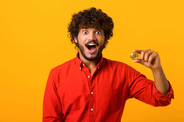 Elinde altın bitcoin kripto para tutan sakallı adam hayretle kameraya bakıyor, fiyattan şok oluyor. Kapalı stüdyo çekimleri turuncu renkte. — Stok fotoğraf