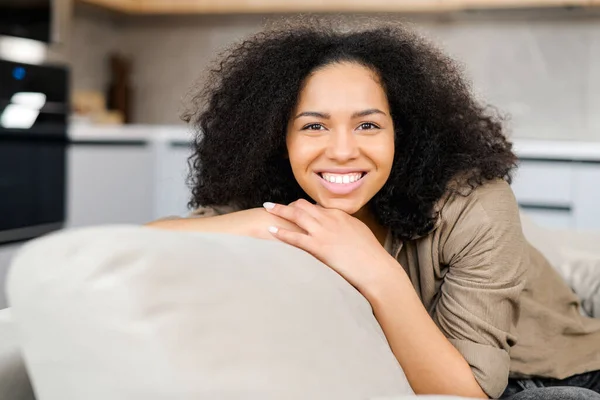 Porträtt av charmig multiracial kvinna med brunett hår. Leende kvinna sitter på soffan och tittar på kameran — Stockfoto