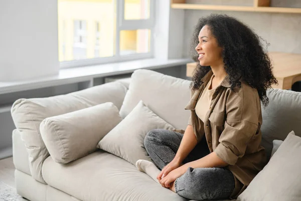 Lugnt sorglös ung kvinna med långt hår sitter vid soffan i modern lägenhet, tittar bort. Kvinna som vilar hemma, dagdrömmer — Stockfoto