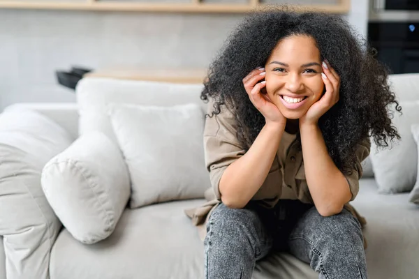 Friska kvinna med brunett hår med glödande ögon, sitter på ren bekväm soffa i modern lägenhet, klädd i avslappnade kläder — Stockfoto