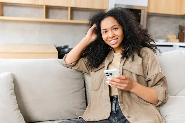 Mujer multiracial mirando a la cámara y sosteniendo el teléfono inteligente en sus manos. Mujer usando el teléfono mientras está sentado en el sofá —  Fotos de Stock