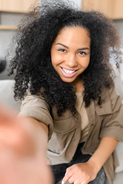 Smiling young multiracial teenage girl sitting on couch in the living room, video-calling via mobile phone, talking to friend, family, taking selfie for social media — Stock Photo, Image