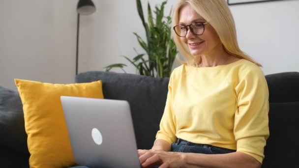 Ανώτερη ελεύθερη επαγγελματίας γυναίκα σε ένα κίτρινο πουλόβερ, κάθεται άνετα στον καναπέ — Αρχείο Βίντεο
