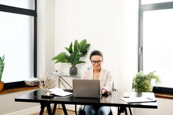 Сосредоточенная и уверенная в себе женщина в очках использует ноутбук для удаленной работы, сидя за столом в офисе. Концентрированная пожилая женщина печатает ответы на письма — стоковое фото