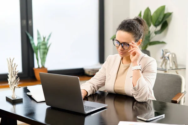 Веселая сотрудница работает с ноутбуком в современном офисе. Положительная женщина средних лет в элегантной повседневной одежде, печатающая на клавиатуре — стоковое фото
