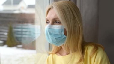 Koruyucu maskeli olgun bir kadın pencereye bakıyor ve üzgün hissediyor.