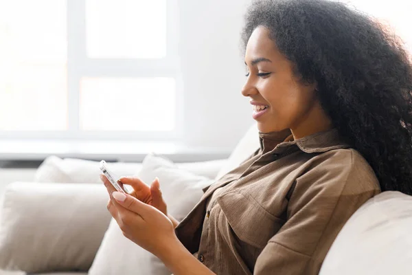 Side view a fiatal multiraciális nő segítségével trendi okostelefon ül a kanapén otthon, etnikai női csevegés a szociális média, SMS-ezés, görgetés hírek feed, megosztani fotók — Stock Fotó