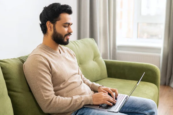 Uomo etnico ottimista in abbigliamento casual con laptop seduto sul divano, giovane studente indiano di sesso maschile che guarda webinar, corsi educativi, apprendimento a distanza — Foto Stock