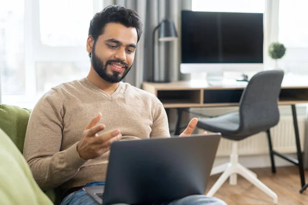 Cheerful middle eastern man in casual wear is sitting on the comfortable sofa and using trendy laptop for video call, talking emotionally with online interlocutor