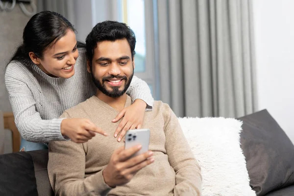 Весела пара проводить час зі смартфоном вдома. Дружина і чоловік дивиться на екран з посмішками, сидячи на дивані в затишній вітальні, дивитися відео — стокове фото