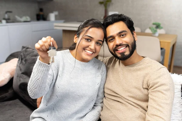 Молода подружня пара весело посміхається і показує ключі від нової квартири, обіймаючись і дивлячись на камеру, стоїть на сучасній кухні нового будинку — стокове фото