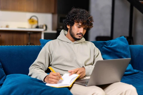 Serious young curly man using laptop computer for studying and work on the distance. Bearded guy watching webinar, taking educational course online — Stock Photo, Image