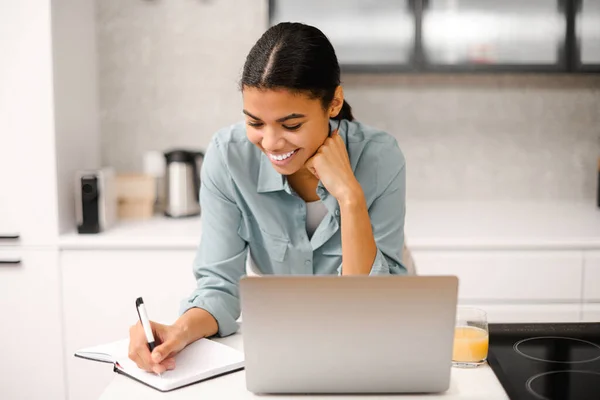 Mujer joven afroamericana está utilizando el ordenador portátil para el trabajo remoto o estudiar en línea, toma notas viendo webinars o clases en la mesa de la cocina —  Fotos de Stock