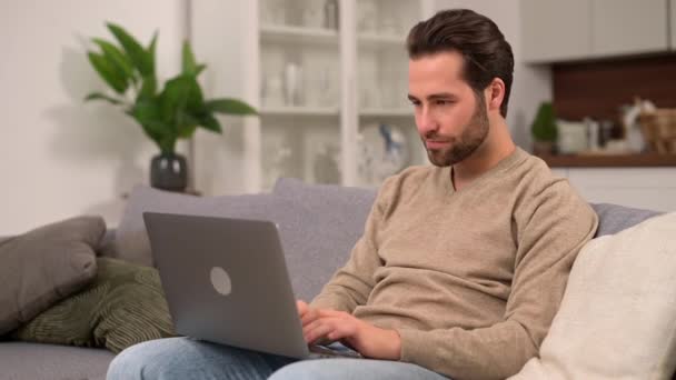 Веселый кавказский мужчина в повседневной одежде сидит на диване и пользуется ноутбуком в современном домашнем офисе — стоковое видео