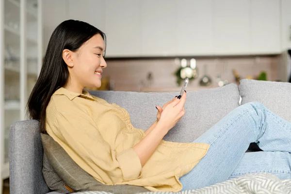 Nyugodt fiatal nő alkalmi viselet feküdt a kanapén a kényelmes nappaliban, és gazdaság okostelefon. Nő időt töltenek online, SMS-t a barátaival — Stock Fotó