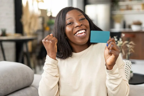 Glad ung kvinna ler med glädje, medan du sitter vid soffan med kreditkort och tittar på kameran. Glad biracial flicka visar plast kreditkort — Stockfoto