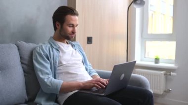 Neşeli beyaz adam modern oturma odasındaki kanepede tek başına oturuyor ve son moda dizüstü bilgisayar kullanıyor.