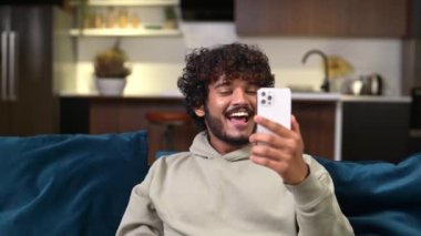 Kanepede oturan heyecanlı genç Hintli adam video bağlantısı için akıllı telefonu kullanıyor.