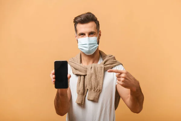 Πορτρέτο του μελαχρινού με ιατρική μάσκα σε λευκό μπλουζάκι που δείχνει το κινητό του και χαμογελά στην κάμερα, συνιστώντας gadget ή mobile εφαρμογή. Εσωτερική στούντιο πυροβόλησε απομονώνονται σε πορτοκαλί — Φωτογραφία Αρχείου