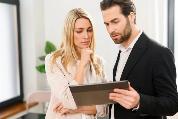 Bella donna e uomo serio con un tablet digitale in mano è in piedi in ufficio, guardando sullo schermo del tablet — Foto Stock