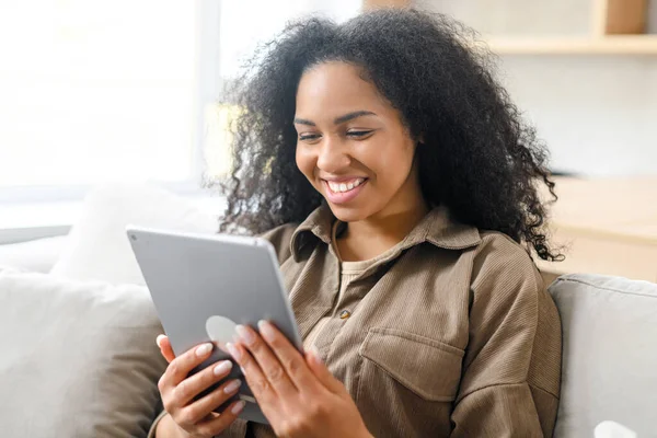 Mladá multiraciální žena s kudrnatými černými vlasy sedí na pohodlném gauči s úsměvem a drží digitální tablet, čtení něco potěšení — Stock fotografie