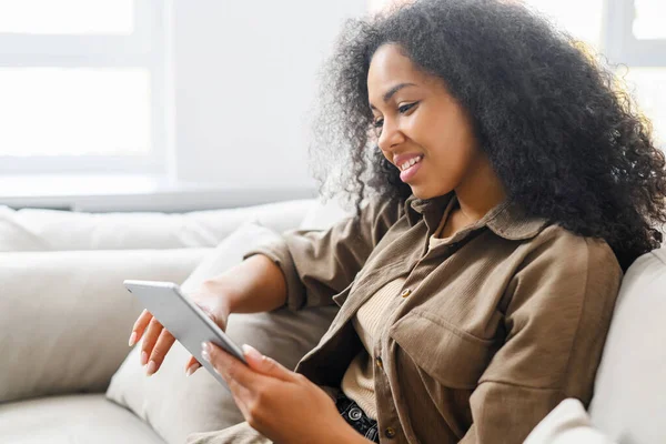 Mladá multiraciální žena s kudrnatými černými vlasy sedí na pohodlném gauči s úsměvem a drží digitální tablet, mluvit s přítelem on-line na videohovoru — Stock fotografie