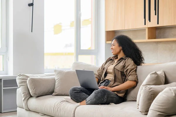 Visão completa da jovem mulher de negócios multirracial ou freelancer feminino em roupas casuais confortáveis sentadas no sofá e trabalhando no projeto, digitando e-mail no laptop — Fotografia de Stock