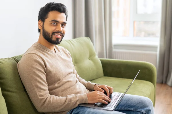 Young Indian male student watching webinars, educational courses, learning on the distance. Front view of positive Hindi man in smart casual clothes using laptop while sitting at the sofa — Stock Photo, Image