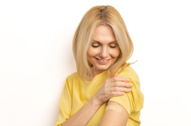 Happy mature lady on white empty blank studio background looking at her hand, smiling and showing arm after vaccine injection. Coronavirus, Covid-19, flu vaccination and immunization