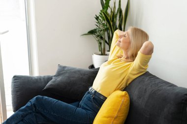 Light-hearted positive middle-aged woman with long hair at the sofa in modern apartment. 50s caucasian female resting at home, looks away lost in dreams