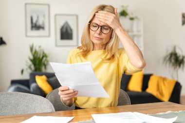 Confident elderly businesswoman is doing paperwork while sitting at the table at the office. Focused senior employee preparing documents, checking bills