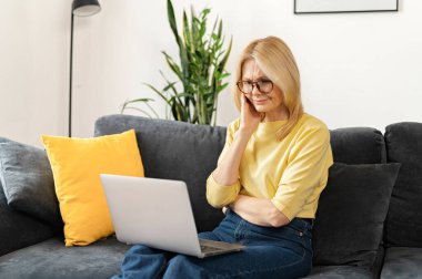 Brooding middle-aged woman using a laptop for remote work at home, studying online, a serious female mature teacher checking tasks