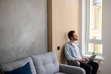 Gündelik giysiler giyen neşeli beyaz adam kanepede oturuyor ve modern evde dizüstü bilgisayar kullanıyor. Klavyede yazan kendinden emin erkek girişimci