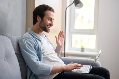 Rahat koltukta oturan neşeli beyaz adam video görüşmesi için son moda dizüstü bilgisayarını kullanıyor, el sallıyor, internetteki interlokasyonu selamlıyor.
