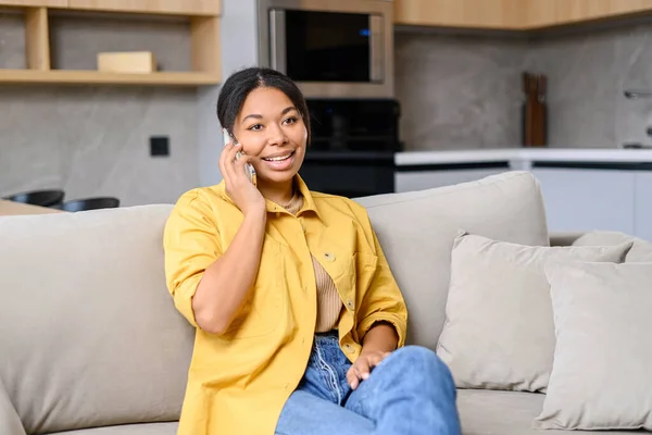 Glad afroamerikansk kvinna pratar via smartphone sittandes på soffan hemma — Stockfoto