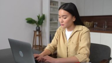 Zeki Asyalı kadın kameraya bakıyor, dizüstü bilgisayarın önünde oturuyor.